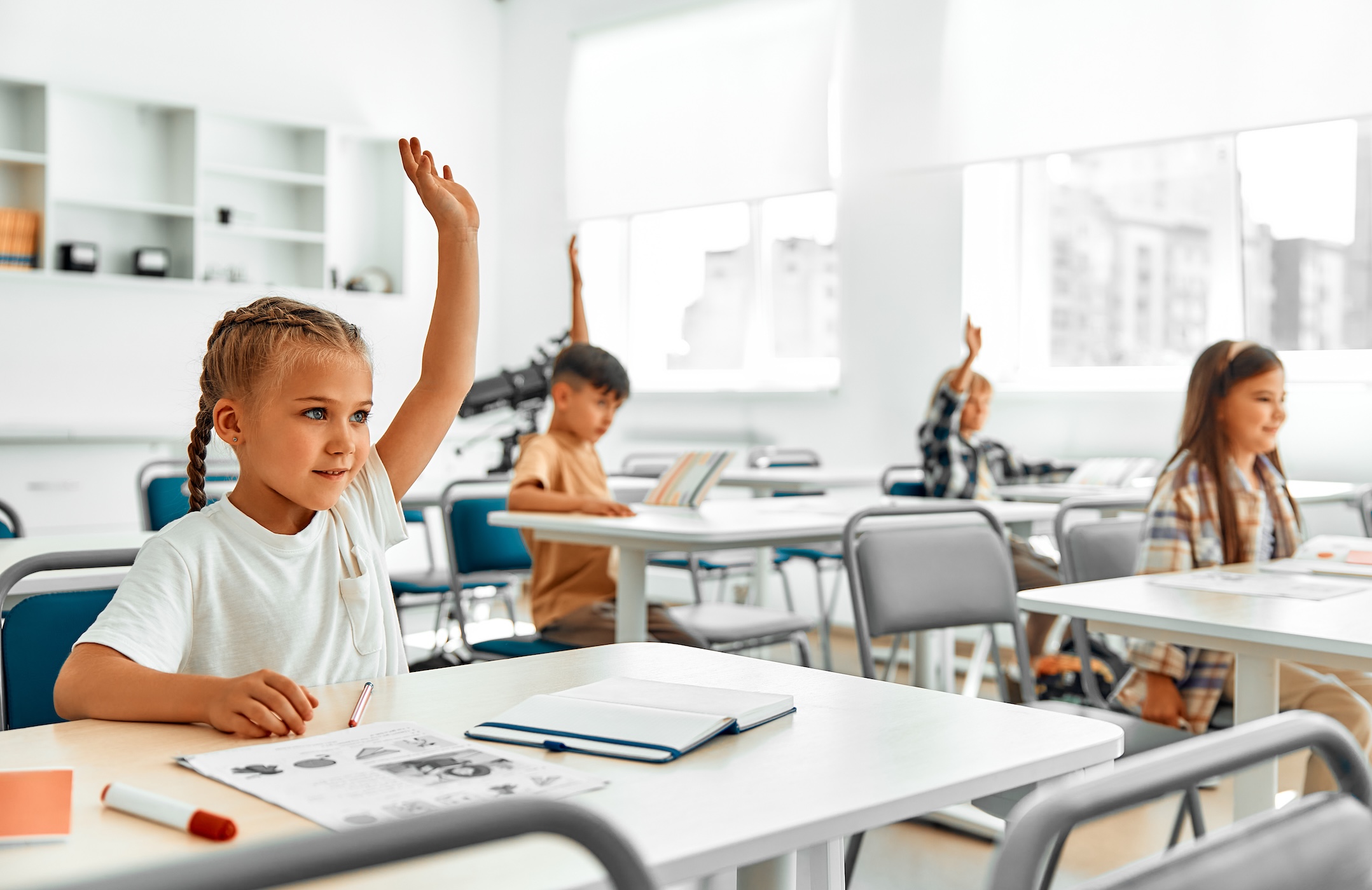 Los alumnos de primer grado en clase, sentados en los escritorios en un aula luminosa, levantando las manos para responder a la pregunta del profesor. Concepto de ciencia y educación. Niños en la escuela.